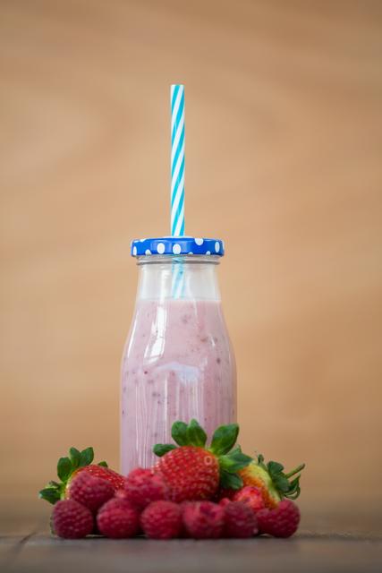 Fresh berry smoothie in a glass bottle with a blue and white striped straw, surrounded by strawberries and raspberries on a wooden surface. Ideal for promoting healthy eating, vegan recipes, summer refreshments, and organic food products. Perfect for use in food blogs, health and wellness websites, and social media posts about nutritious drinks.