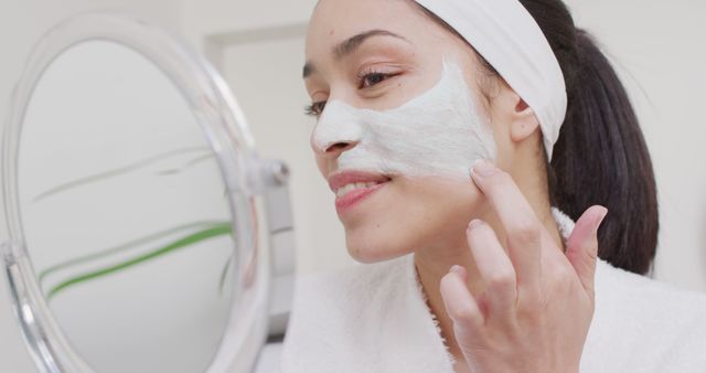 Young woman is applying a face mask cream as part of her daily skincare routine. She is smiling and looking into a round mirror. Ideal for use in beauty blogs, skincare product promotions, and wellness articles.
