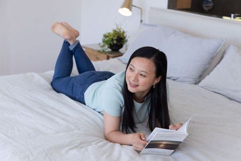 Mature asian woman relaxing on bed reading paperback in serene minimalist pastel bedroom