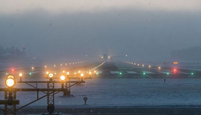 Commercial airliner approaching snowy runway through foggy twilight with lit approach lights