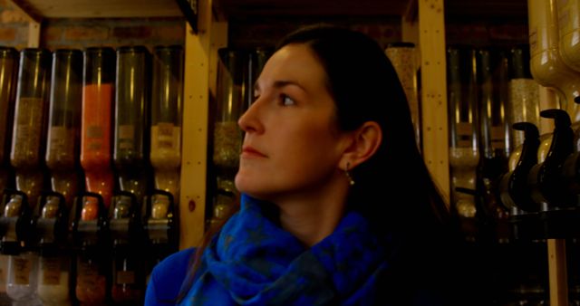 A woman with dark hair wears a blue scarf while standing in a bulk food store, looking off to the side. Behind her are shelves filled with various grains and ingredients stored in jars. This image can be used for themes related to sustainability, eco-friendly shopping, and zero-waste lifestyles.