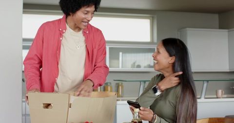 Young couple unpacking moving box in modern kitchen laughing while using smartphone
