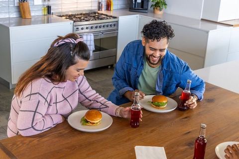 Couple laughing and sharing burgers at kitchen island with red sodas, cozy casual meal