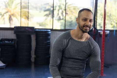 Mid-adult man sitting on bench smiling after workout in bright gym with palm trees