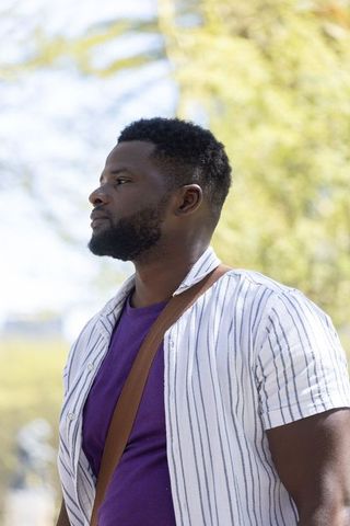 African american man standing in sunlit park wearing purple tee and striped shirt
