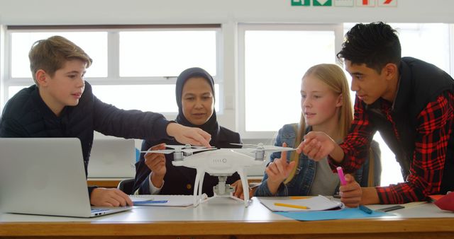 This image showcases a diverse group of teenage students working together with their teacher to explore drone technology in a STEM education activity. They are all engrossed in examining the features and functionality of the drone, which fosters an environment of curiosity, collaboration, and hands-on learning. Ideal for use in educational websites, technology blogs, and promotional materials highlighting innovative teaching methods, teamwork in education, and the integration of modern technology in classrooms.
