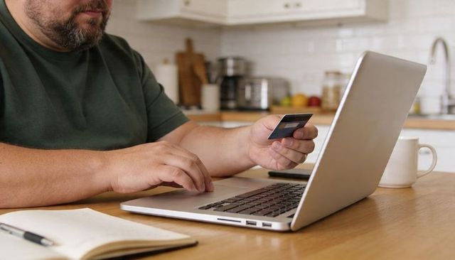 Man using laptop and holding credit card for online payment in modern home kitchen