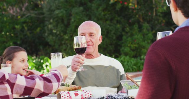 Family gathering for celebration, sharing toast with wine outdoors. Elderly man raising wine glass surrounded by family members at table enjoying festive meal. Good for family events, celebrations, festivals, holiday adverts, promoting outdoor activities, family unity concepts.