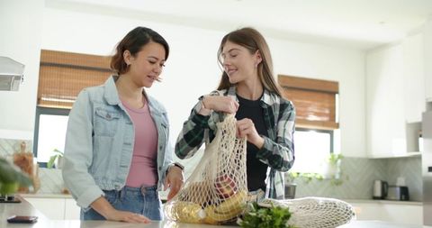 Friends unpacking reusable produce bag on bright kitchen island, smiling over fresh fruit
