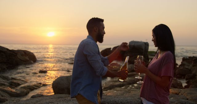 Couple standing by the sea at sunset, holding wine glasses, and sharing a romantic moment. Man in blue shirt pouring wine for woman in pink top. Suitable for themes related to romance, relaxation, vacations, leisure activities, and joyous moments.