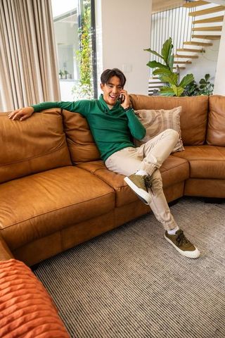 Asian man sitting on tan leather sofa talking on smartphone in contemporary home