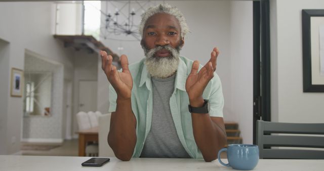 Mature man with gray hair and beard talks at his kitchen table, with a coffee mug and smartphone in view. Uses for home lifestyle blogs, remote work articles, senior engagement content, and casual home setting promotions.