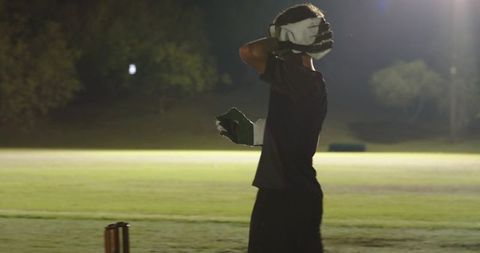 Nighttime cricketer adjusting helmet and gloves by stumps on floodlit pitch preparing to bat