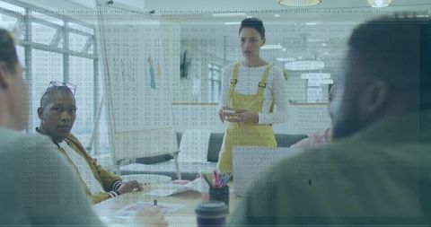 Woman Presenting in Yellow Overalls Leading Team Meeting in Modern Glass Boardroom