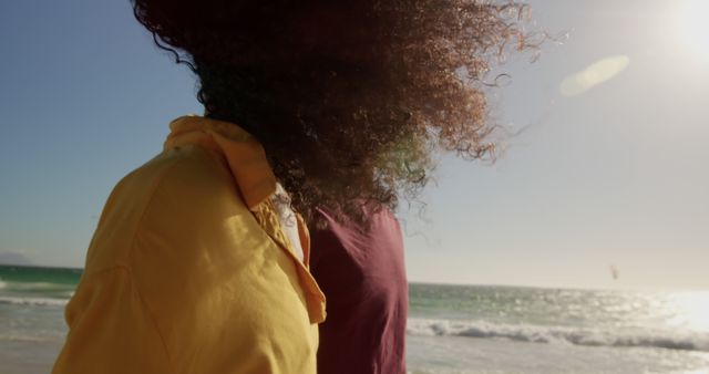 Couple enjoying sunny beach day together with curly hair blowing in wind. Red and yellow shirts create colorful contrast against blue ocean and sky. Ideal for travel promotions, lifestyle blogs, vacation themes, and ads focusing on relaxation and happy moments.