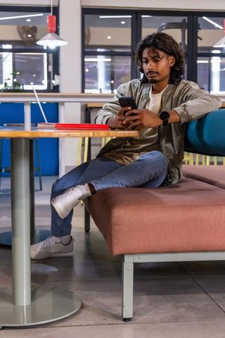 Indian young man using smartphone while relaxing on cafe bench in modern coworking space