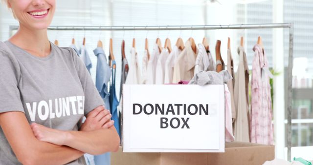 A young Caucasian woman smiles while standing next to a donation box in a clothing drive setting, with copy space. Her volunteer shirt suggests a charitable event, emphasizing the importance of giving to those in need.