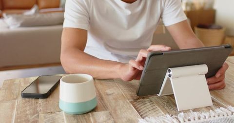 African american man using tablet at wooden table with smartphone and coffee mug, home workspace
