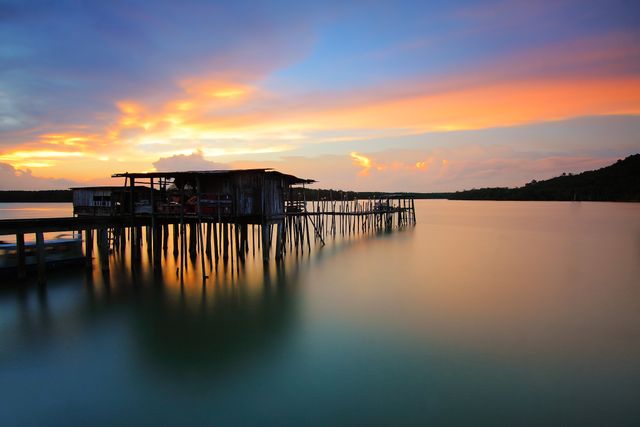This image captures a rustic wooden pier extending over calm waters during a vibrant sunset. The colorful sky and the stillness of the water create a peaceful and tranquil scene. Perfect for use in travel blogs, websites promoting serene getaways, or inspirational nature-themed content. Can be used to emphasize tranquility, relaxation, and natural beauty.