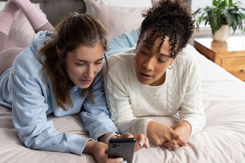 Cozy morning browsing: diverse female friends lying on bed sharing smartphone together