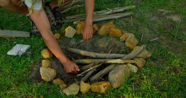 Person preparing a campfire pit with stones and sticks in a forest clearing. Suitable for outdoor and camping-related themes, wilderness survival guides, and recreational activity promotions.
