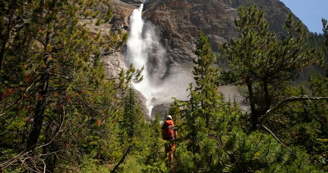 Person hiking through forested area towards large waterfall cascading down rocky cliff. Ideal for promoting outdoor activities, nature travel destinations, and adventure tours. Perfect for websites, blogs, or publications focused on hiking, trekking, and exploring pristine natural landscapes.