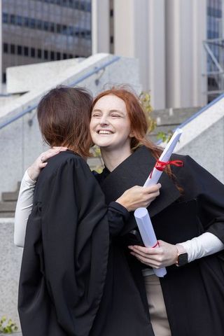 Smiling female graduates hugging, holding red-ribbon diplomas on urban campus steps