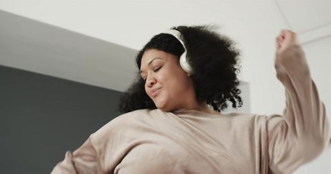 Joyful woman dancing at home wearing white over-ear headphones and cozy beige sweatshirt