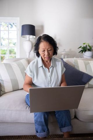 Senior asian woman using laptop on sofa in bright cozy living room