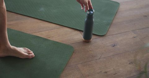 Reaching for insulated bottle during home yoga session on green mats and wood floor