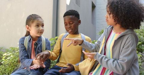 Diverse elementary children playing hand game on school bench with backpacks
