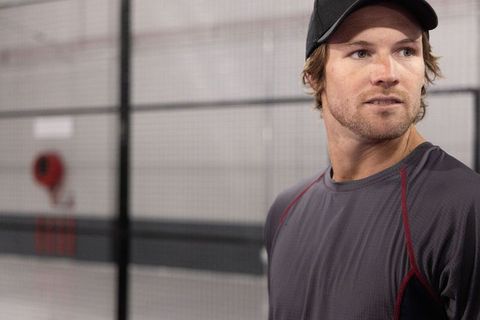 Late-twenties athlete standing by gym cage wearing cap and performance shirt, copy space