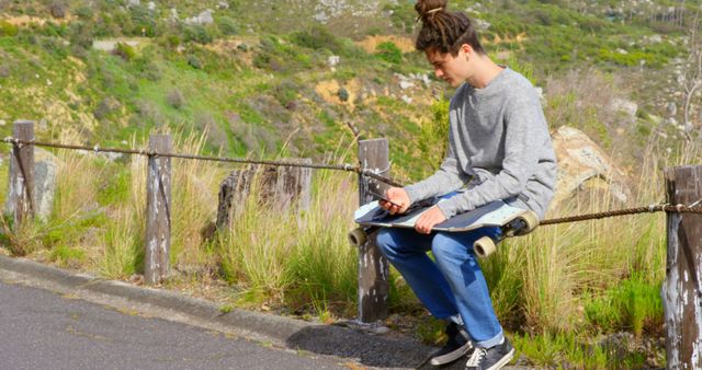 Young teenage boy sits on a rustic wooden fence with his skateboard outdoors. He is wearing casual clothing and appears to be deep in thought. The background includes green hills and a rural setting, creating a serene and reflective mood. Ideal for themes like youth lifestyle, outdoor activities, relaxation, and nature.