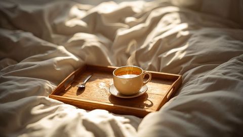 Sunlit bed tray with steaming coffee cup on rumpled white comforter morning calm cozy