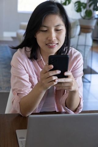 Asian Woman Using Smartphone While Working on Laptop at Home