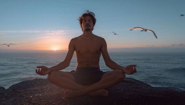 Shirtless man meditating on coastal cliff at sunset with seagulls and ocean horizon, mindfulness
