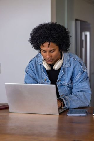 African american woman typing on laptop wearing denim jacket headphones in home workspace