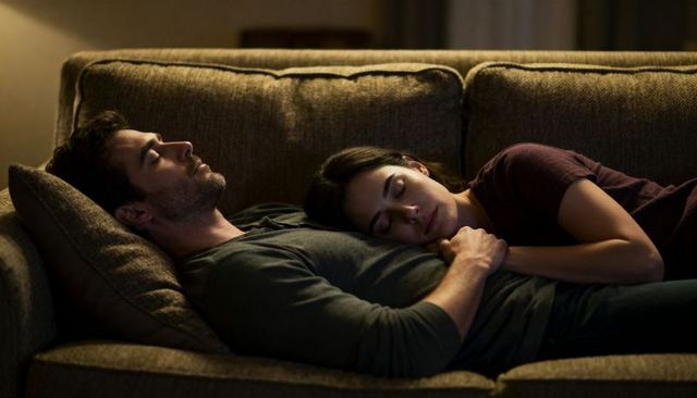 Couple cuddling and dozing on brown sofa under warm lamplight, wearing grey shirt and maroon top