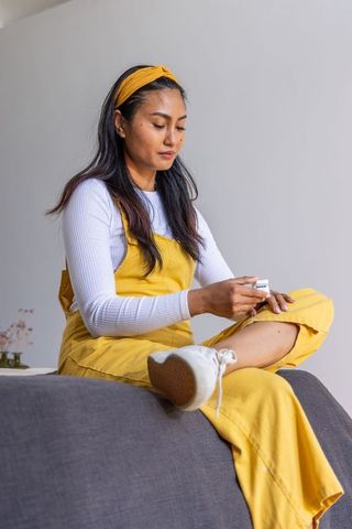 Asian woman applying moisturizer to lower leg wearing yellow overalls and headband