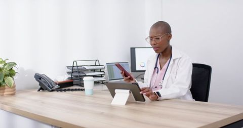 Clinician managing telehealth on tablet and phone in modern minimalist clinic workspace