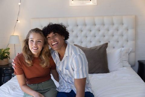 Smiling mixed-race couple cuddling on bed in cozy bedroom with tufted headboard and string lights