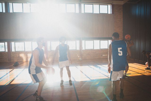 Diverse male basketball team practicing on an indoor court with sunlight streaming through windows. Ideal for use in sports-related content, teamwork and training articles, fitness and exercise promotions, and athletic event advertisements.