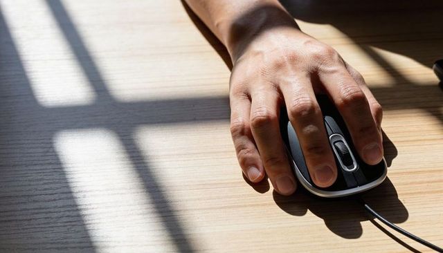 Right hand resting on wired mouse with window shadow on wood desk, closeup detail and cable