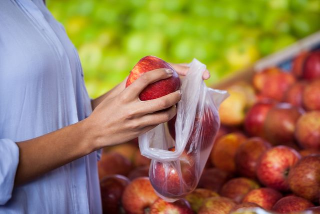Woman selecting fresh apples in a supermarket, placing them in a plastic bag. Ideal for use in articles or advertisements related to healthy eating, grocery shopping, fresh produce, and lifestyle choices. Can also be used in content promoting organic food, nutrition, and market shopping experiences.
