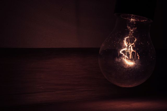 This image showcases a dimly lit vintage light bulb with its filament glowing against a dark wood background. The atmosphere portrays a classic and rustic feel, ideal for use in illustrating concepts of energy, electricity, or vintage interior design. Can be used in blogs, articles, advertisements, or websites focusing on lighting, technology, or home decor.