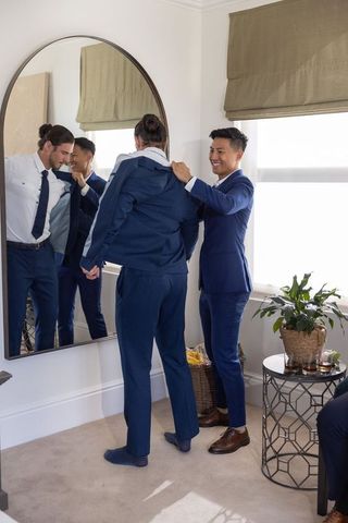 Diverse male friends pulling navy suit jacket while smiling in bright dressing room mirror