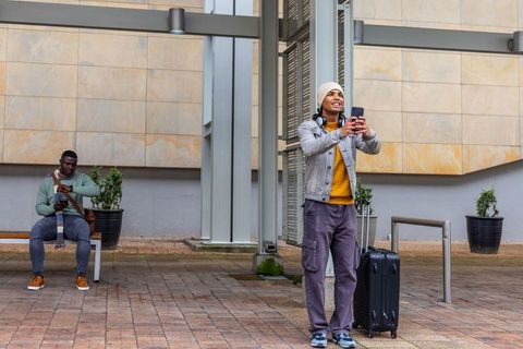 Taking selfie at covered urban transit plaza with rolling suitcase and waiting commuter