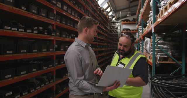 Two warehouse staff talking while holding a laptop in a well-organized storage aisle filled with shelves and boxes. Perfect for using in promotional materials for logistics companies, safety protocols, teamwork importance, and warehouse management software advertisements.