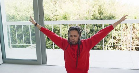 Meditating and stretching man kneeling by sunlit balcony wearing red hoodie