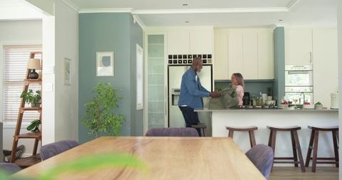 Multicultural couple unpacking grocery bags at modern kitchen island with cozy dining table